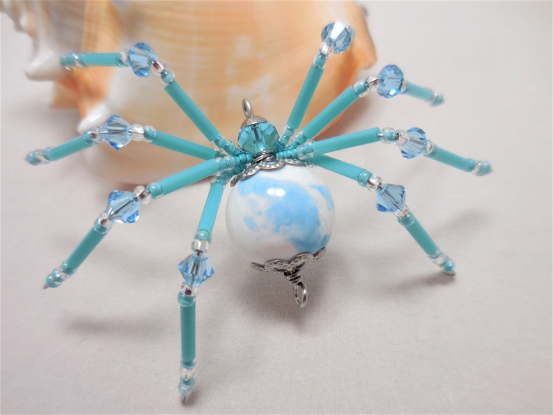 Puede incluir: Un amuleto de ara&ntilde;a de cuentas con un cuerpo esf&eacute;rico de m&aacute;rmol azul y blanco, patas turquesa y detalles de cristal transparente.