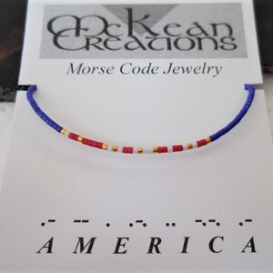 May include: A blue, red, white, and gold beaded bracelet with the word "AMERICA" spelled out in Morse code below. The bracelet is on a white card with the words "McKean Creations Morse Code Jewelry".
