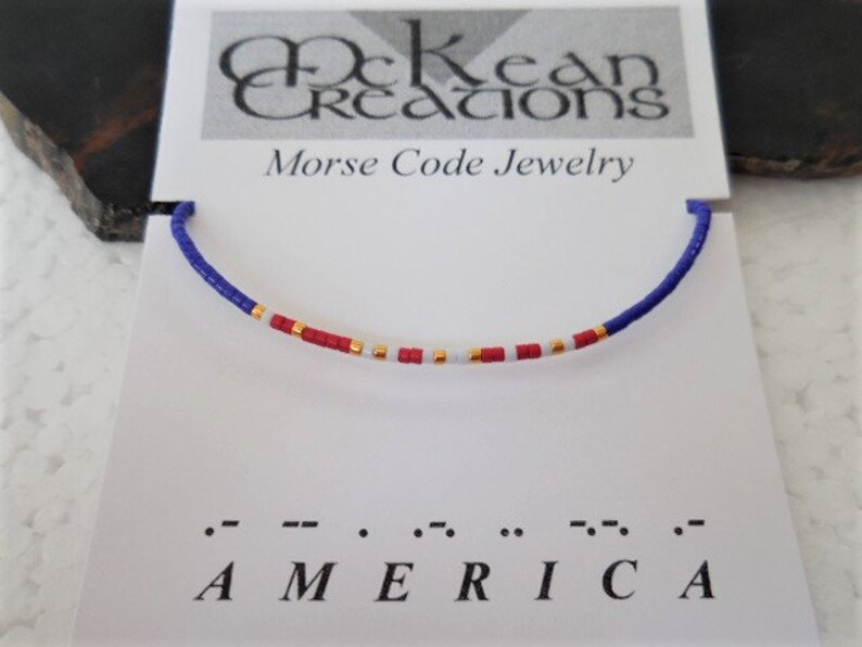 May include: A blue, red, white, and gold beaded bracelet with the word "AMERICA" spelled out in Morse code below. The bracelet is on a white card with the words "McKean Creations Morse Code Jewelry".