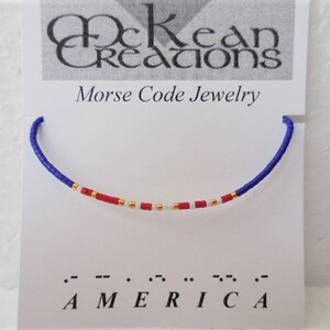 May include: A blue, white, and red beaded bracelet with gold beads spelling out "AMERICA" in Morse code. The bracelet is displayed on a white card with the text "McKean Creations Morse Code Jewelry".