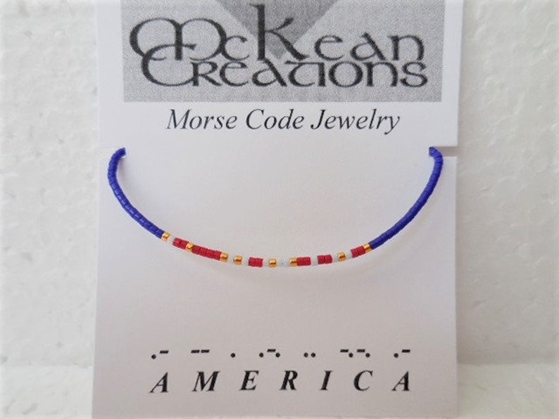 May include: A blue, white, and red beaded bracelet with gold beads spelling out "AMERICA" in Morse code. The bracelet is displayed on a white card with the text "McKean Creations Morse Code Jewelry".