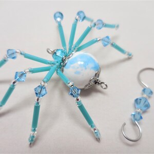 Puede incluir: Un adorno de ara&ntilde;a de cuentas turquesa con un cuerpo esf&eacute;rico de m&aacute;rmol blanco y azul y detalles plateados. La ara&ntilde;a tiene ocho patas y est&aacute; unida a un gancho plateado.