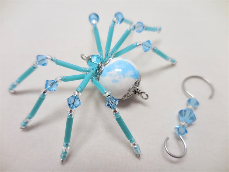Puede incluir: Un adorno de ara&ntilde;a de cuentas turquesa con un cuerpo esf&eacute;rico de m&aacute;rmol blanco y azul y detalles plateados. La ara&ntilde;a tiene ocho patas y est&aacute; unida a un gancho plateado.