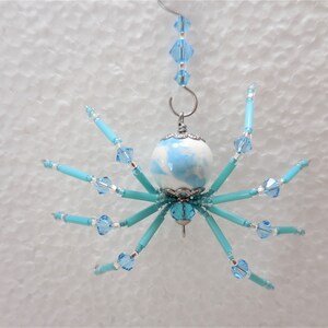 Puede incluir: Un adorno de ara&ntilde;a azul y blanco con un cuerpo y patas de cuentas. La ara&ntilde;a est&aacute; colgada de un gancho plateado.