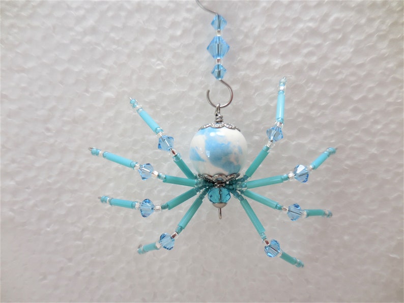 Puede incluir: Un adorno de ara&ntilde;a azul y blanco con un cuerpo y patas de cuentas. La ara&ntilde;a est&aacute; colgada de un gancho plateado.