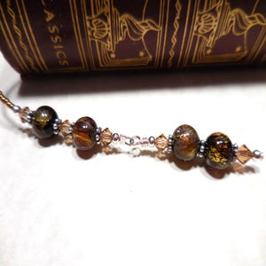 May include: A beaded bookmark with brown and gold beads and silver accents. The bookmark features a series of round beads with a marbled effect, interspersed with smaller, faceted beads. The bookmark is placed in a book.