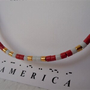 May include: A red, white, and gold beaded bracelet with the word "AMERICA" spelled out in braille below it.