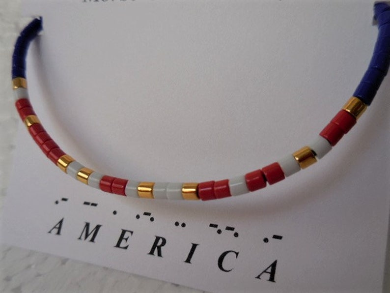 May include: A red, white, and gold beaded bracelet with the word "AMERICA" spelled out in braille below it.
