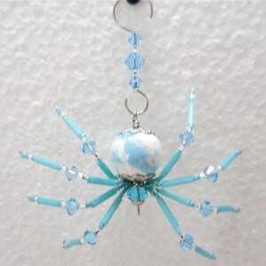 Puede incluir: Un adorno de ara&ntilde;a azul y blanco con un cuerpo de cuentas de cristal y patas de cuentas azules. La ara&ntilde;a est&aacute; colgada de un gancho plateado con una cadena de cuentas de cristal.