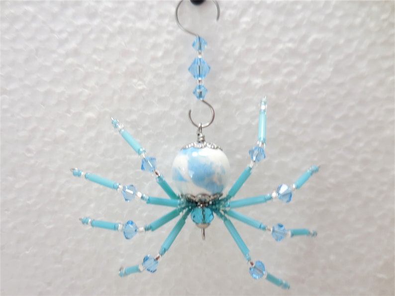 Puede incluir: Un adorno de ara&ntilde;a azul y blanco con un cuerpo de cuentas de cristal y patas de cuentas azules. La ara&ntilde;a est&aacute; colgada de un gancho plateado con una cadena de cuentas de cristal.