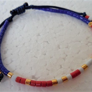 May include: A blue, white, and red beaded bracelet with gold accents. The bracelet is made with small, round beads and has a knotted closure.
