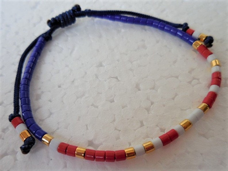May include: A blue, white, and red beaded bracelet with gold accents. The bracelet is made with small, round beads and has a knotted closure.