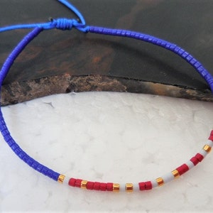 May include: A blue, red, and white beaded bracelet with gold accents. The bracelet is made of small, square beads and is tied with a blue cord.