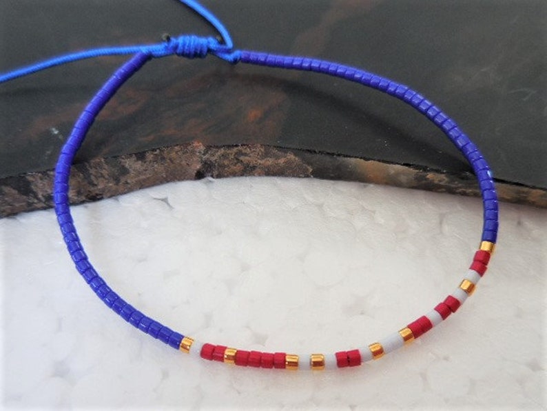 May include: A blue, red, and white beaded bracelet with gold accents. The bracelet is made of small, square beads and is tied with a blue cord.