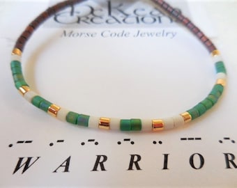 May include: A green, white, and gold beaded bracelet with the word "WARRIOR" spelled out in Morse code below. The bracelet is made with small, round beads and has a brown cord.