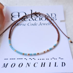 May include: A brown leather cord bracelet with blue and white beads and gold accents. The bracelet is tied with a sliding knot. The bracelet is displayed on a white card with the text "Ocean Creations Morse Code Jewelry" and the Morse code for "MOONCHILD".