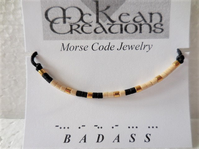 May include: A black string bracelet with white and gold beads spelling out "BADASS" in Morse code. The bracelet is on a white card with the text "McKean Creations Morse Code Jewelry".