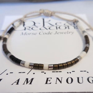 May include: A brown, white, and silver beaded bracelet with a beige cord closure. The bracelet spells out "I AM ENOUGH" in Morse code.