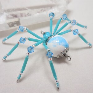 Puede incluir: Una ara&ntilde;a de cuentas turquesa con un cuerpo esf&eacute;rico de m&aacute;rmol blanco y azul y detalles plateados. La ara&ntilde;a tiene ocho patas y est&aacute; decorada con cristales azules transparentes.