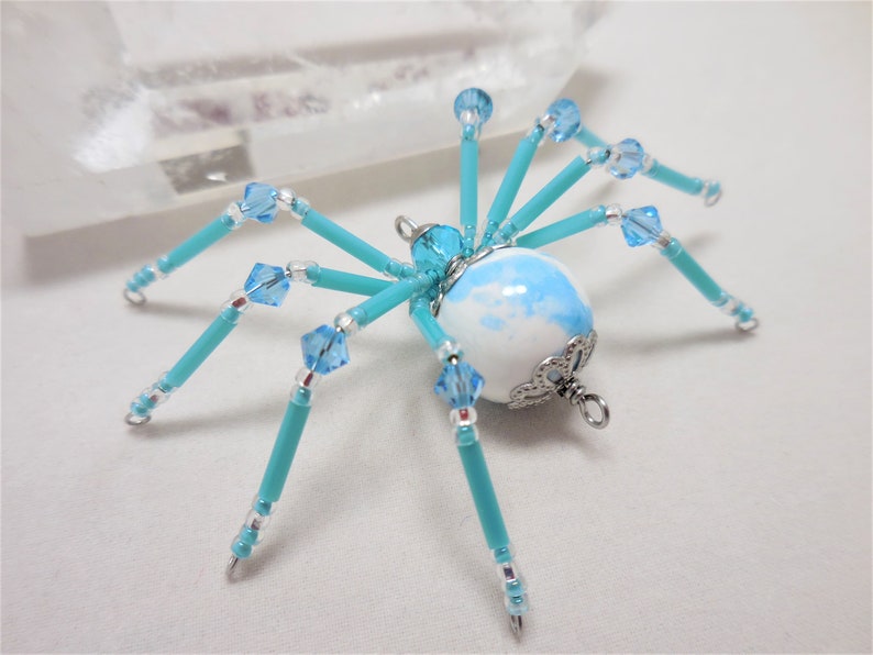 Puede incluir: Una ara&ntilde;a de cuentas turquesa con un cuerpo esf&eacute;rico de m&aacute;rmol blanco y azul y detalles plateados. La ara&ntilde;a tiene ocho patas y est&aacute; decorada con cristales azules transparentes.