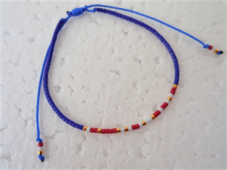 May include: A blue and white beaded bracelet with red accents. The bracelet is made with small, round beads and has a simple knot closure.