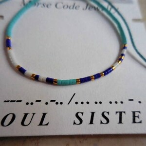 May include: A turquoise, white, gold, and blue beaded bracelet with a white card that reads "SOUL SISTER" in black text and a morse code message.
