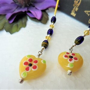 May include: Two yellow heart-shaped glass beads with a flower design in red, white, and green. The beads are attached to a silver chain with blue and gold beads.