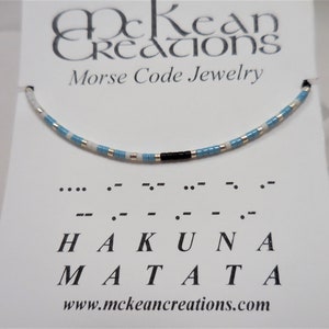 May include: A blue and white beaded bracelet with a black bead accent. The bracelet spells out "Hakuna Matata" in Morse code.