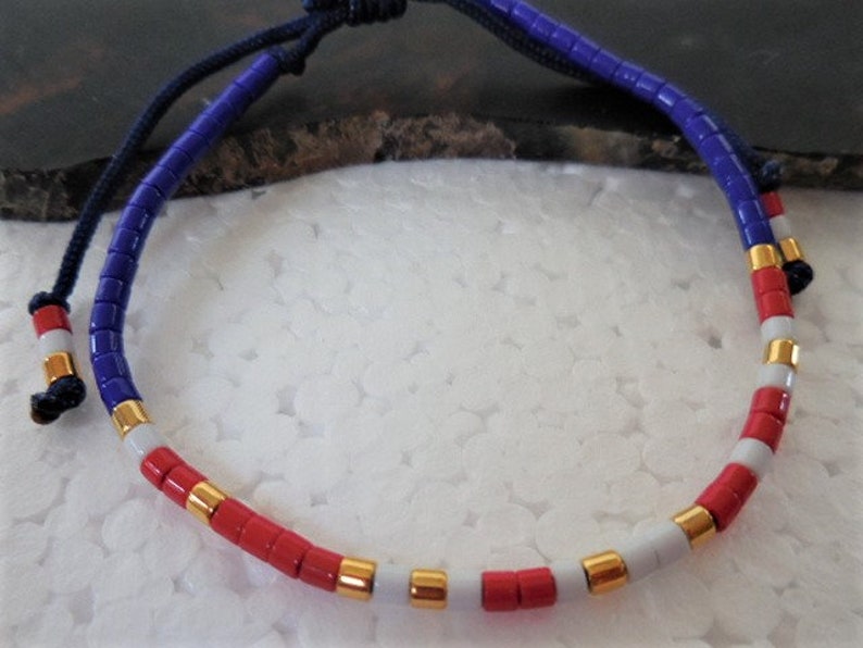 May include: A blue, white, and red beaded bracelet with gold accents. The bracelet is made of small, round beads and has a simple, elegant design.