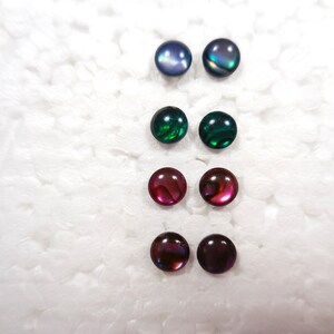 May include: Six pairs of iridescent stud earrings in various colors: blue, green, red, and purple. The earrings have a round, polished design, and are displayed on a white, textured surface. These earrings are a jewelry accessory.