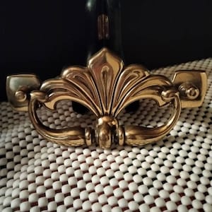 May include: A gold-toned metal cabinet pull with a decorative floral design. The pull has a curved shape and two small, rounded ends.