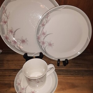 Pode incluir: Um conjunto de três pratos de cerâmica branca com um design floral em tons de rosa e verde. Os pratos têm uma borda cinza e estão dispostos em forma de pirâmide.