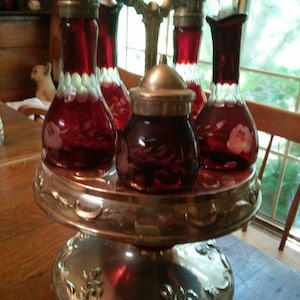 May include: A set of six red glass bottles with white floral designs, on a silver metal stand with a round base. The bottles have silver stoppers and are arranged in a circle on the stand.