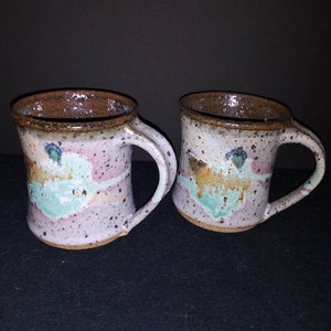 May include: Two handmade ceramic mugs with a speckled glaze in shades of brown, pink, and green. The mugs have a rustic, handmade look.