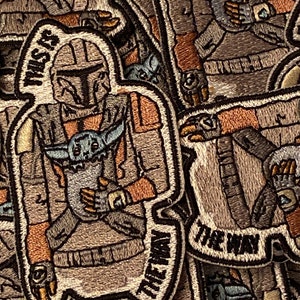 Puede incluir: Una pila de parches bordados con un personaje de dibujos animados de Mandalorian sosteniendo un bebé Yoda. Los parches son beige, marrón y gris con contornos negros. El texto en los parches dice "This is the way".