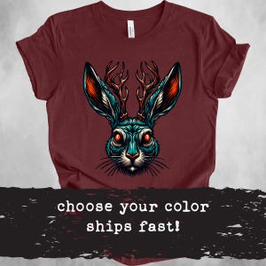 May include: A maroon t-shirt with a graphic of a blue rabbit with antlers. The rabbit has red eyes and a white nose. The text "choose your color ships fast!" is at the bottom of the image.