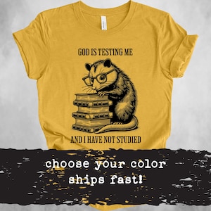 Puede incluir: Una camiseta amarilla con una ilustración en blanco y negro de una zarigüeya con gafas y sosteniendo una pila de libros. El texto "Dios me está poniendo a prueba y no he estudiado" está impreso en la camiseta.