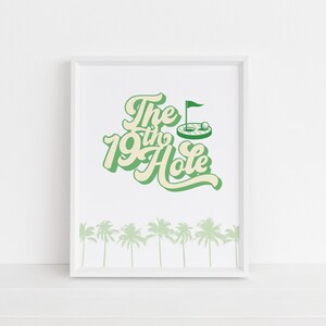 May include: A white framed print with green text that reads "The 19th Hole" with a green golf hole and flag illustration. The bottom of the print has a row of green palm trees.