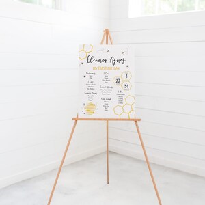 May include: A white poster with a yellow honeycomb pattern and black bee illustrations. The poster has the text "Eleanor Agnes My First Bee Day" and lists information about the baby, such as their name, birthdate, weight, and favorite foods.