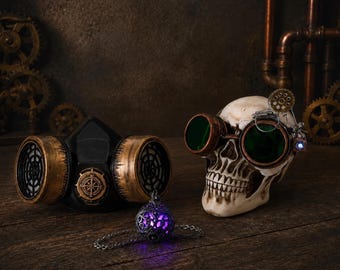 May include: A black respirator mask with metallic gold accents, a human skull wearing copper-rimmed goggles with green lenses, and a small silver-toned pendant with a glowing purple light, all arranged together in a thematic display.