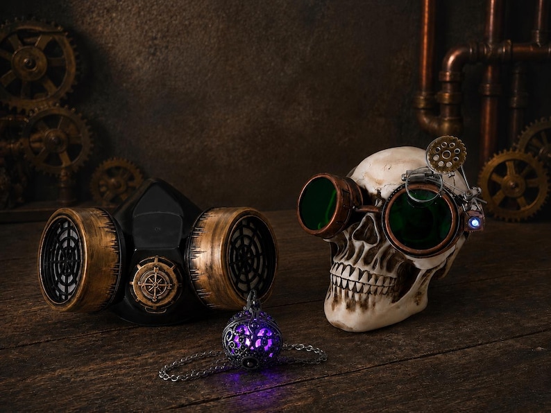 May include: A black respirator mask with metallic gold accents, a human skull wearing copper-rimmed goggles with green lenses, and a small silver-toned pendant with a glowing purple light, all arranged together in a thematic display.