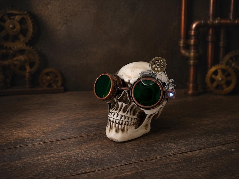 May include: A human skull replica wearing copper-toned goggles with dark green lenses. The goggles feature an attached mechanical gear mechanism and a small magnifying lens on the side, resting over the eye sockets of the off-white bone-colored skull.