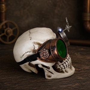 May include: A human skull model wearing copper-colored goggles with green lenses and a small compass detail on the side. A metal band with a small blue LED light is attached to the top of the goggles, secured by a black elastic strap around the skull.