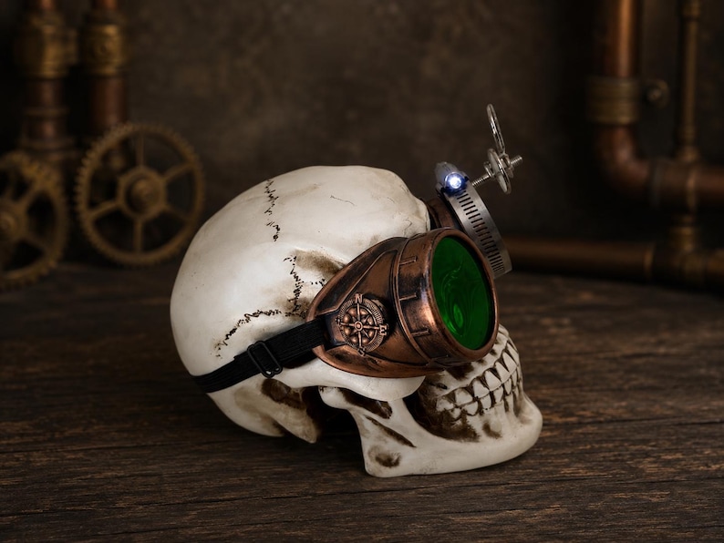 May include: A human skull model wearing copper-colored goggles with green lenses and a small compass detail on the side. A metal band with a small blue LED light is attached to the top of the goggles, secured by a black elastic strap around the skull.