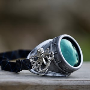 Könnte beinhalten: Silberne Steampunk-Brille mit grünem Glas und einem Oktopus-Anhänger. Die Brille hat ein schwarzes Gummiband.