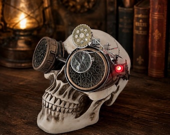 May include: A human skull model wearing metallic goggles with intricate filigree patterns, a small magnifying lens, and a glowing red light on the side. The goggles feature bronze and silver tones, with a gear-like detail on the top frame.