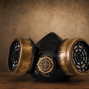 May include: A black respirator mask featuring two large cylindrical filters with a distressed metallic gold finish. The center of the mask displays a circular emblem resembling a compass with cardinal directions N, S, E, and W embossed on it.