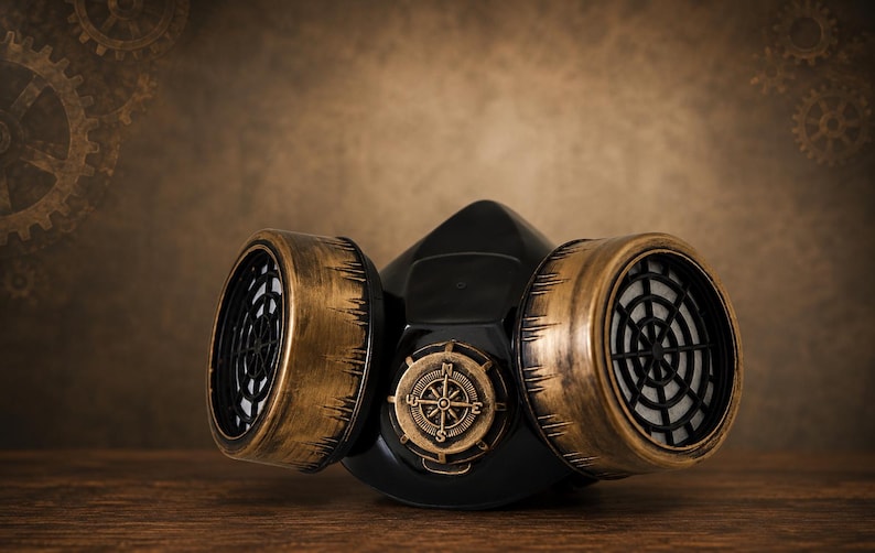 May include: A black respirator mask featuring two large cylindrical filters with a distressed metallic gold finish. The center of the mask displays a circular emblem resembling a compass with cardinal directions N, S, E, and W embossed on it.