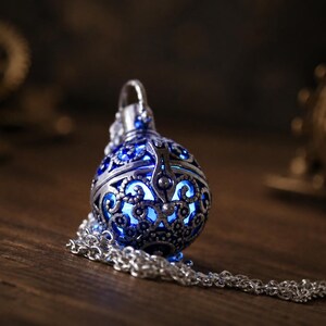 May include: A spherical silver-toned metal pendant featuring intricate filigree cutouts that reveal a glowing blue light from within. The pendant is attached to a silver-toned metal chain that rests in a coiled loop beside the spherical charm.