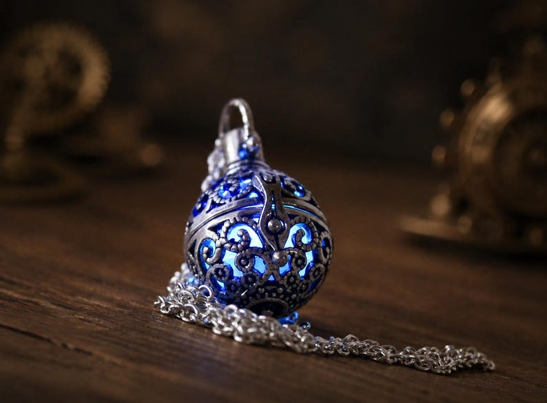 May include: A spherical silver-toned metal pendant featuring intricate filigree cutouts that reveal a glowing blue light from within. The pendant is attached to a silver-toned metal chain that rests in a coiled loop beside the spherical charm.